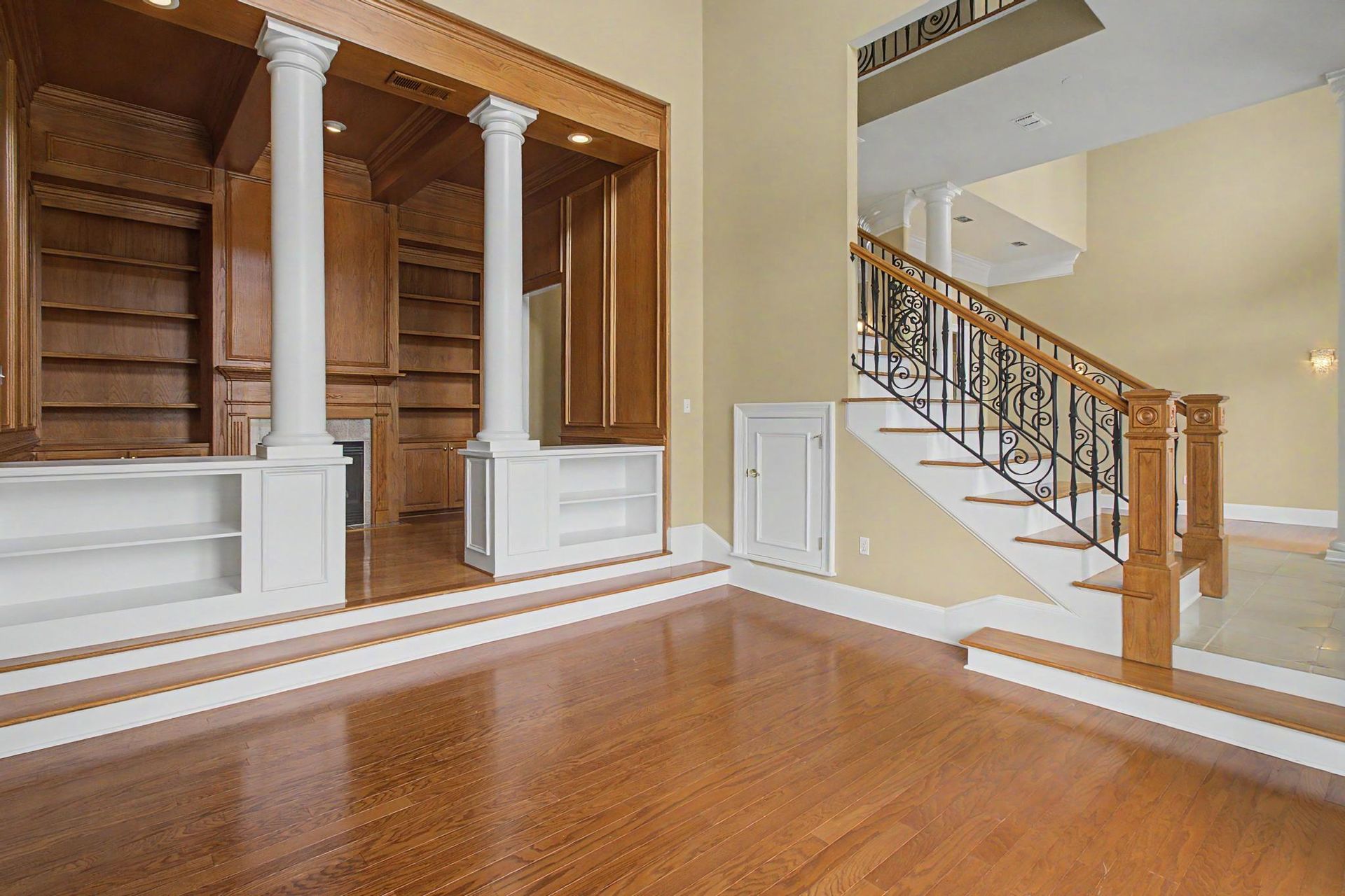 Paneled library or study with floor-to-ceiling oak built-ins, white columns flanking a stepped reading nook, and a wrought-iron staircase visible beyond